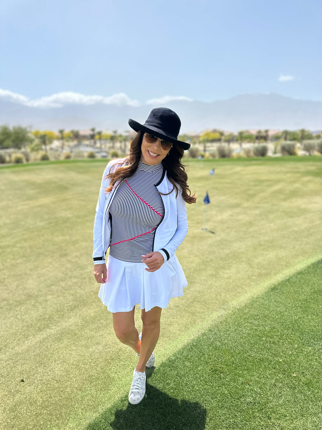 Woman wearing hat, stylish striped black and white shirt, white cardigan and white pleated skort on golf course