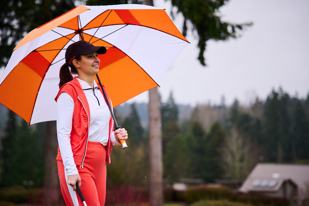 A woman caring an umbrella wearing a coral red vest and pant with a white longsleeve zip front top on the golf course. 