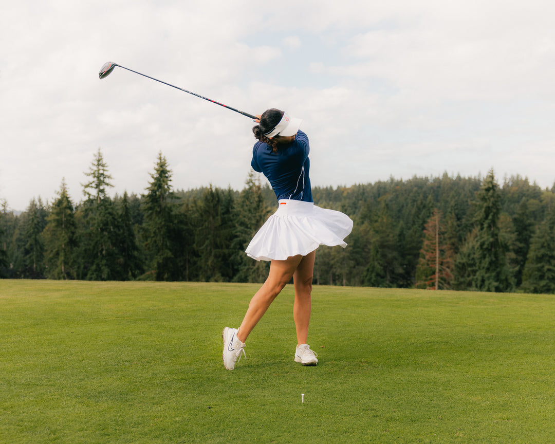 Woman swinging golf club at the course in Navy longsleeve top and white skort