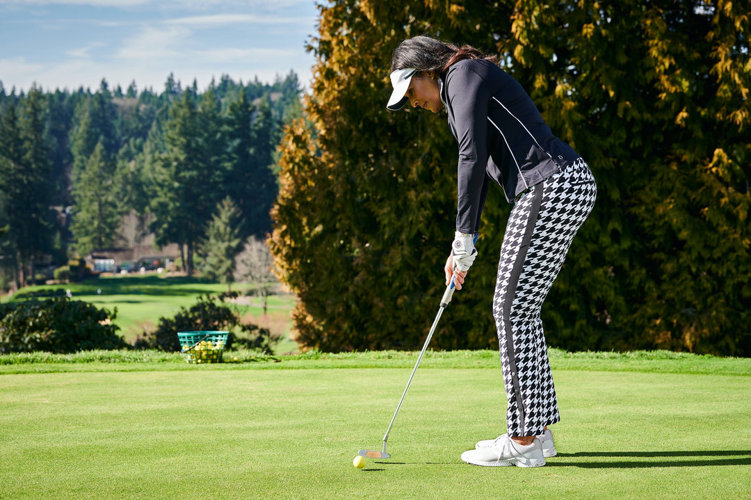 A female golfer putting on the green in KINONA buffalo check golf pants and visor.