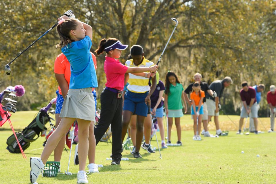 A group of young female golfers lined up practicing on driving range in colorful clothes.