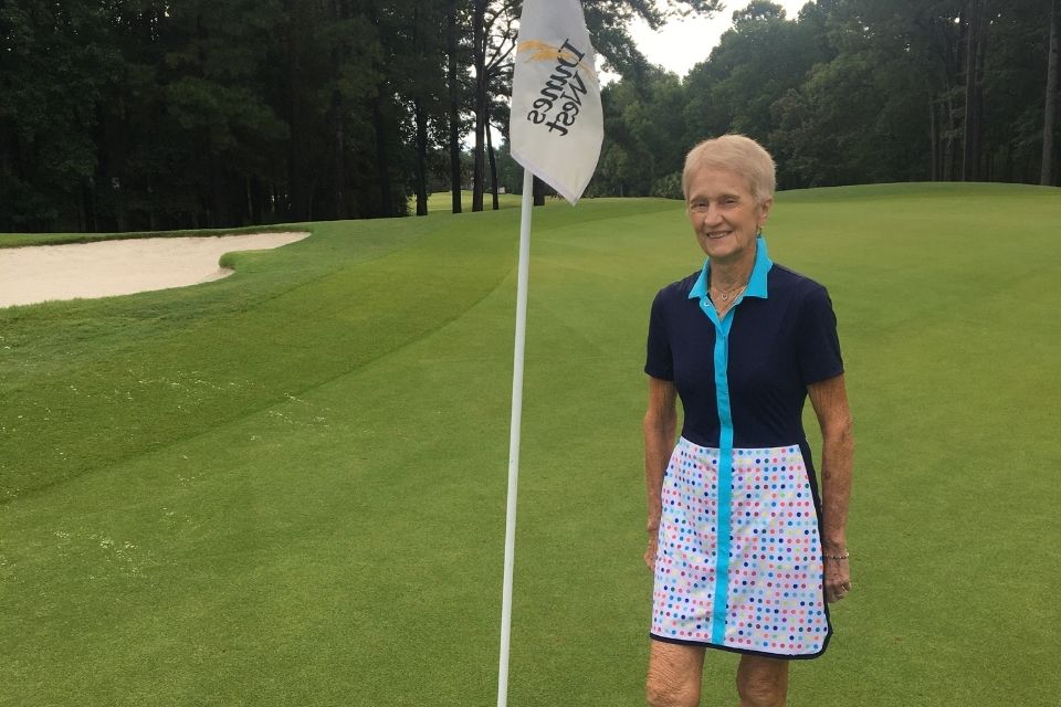 Hilda smiling next to a tee marker on a green grass course in KINONA blue top and white skort. 