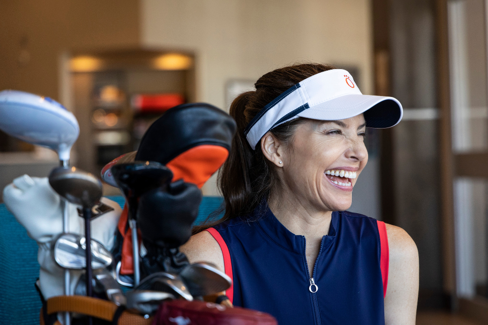 Woman smiling in a white KINONA visor and blue sleeveless golf dress with a set of clubs.