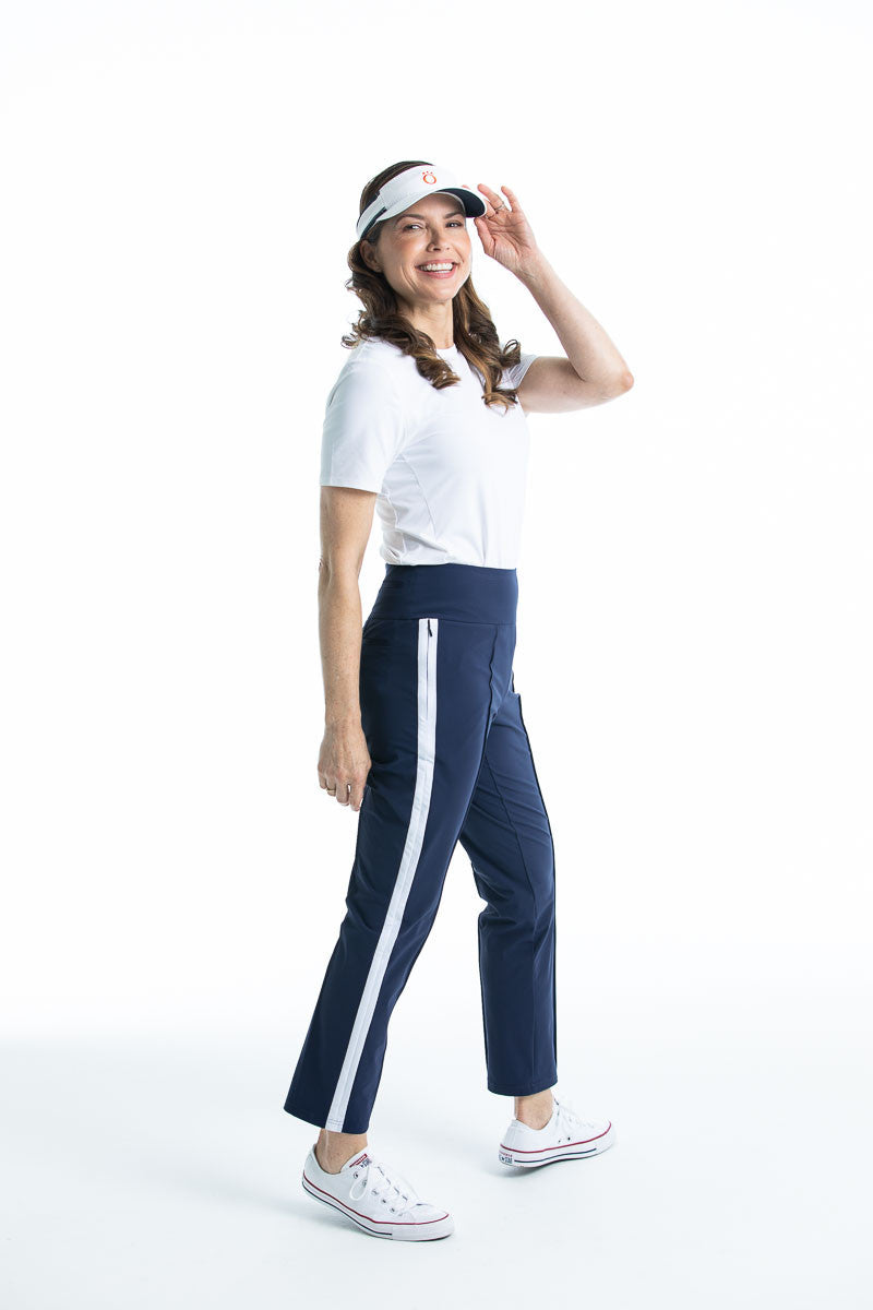 Woman holding the brim of her white No Hat Hair visor wearing a white Tee it Up shortsleeve golf shirt and navy blue Tailored Track golf pant.