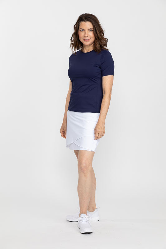Full front view of a woman wearing a Tee It Up Short Sleeve Golf Shirt in navy blue and a Wrap It Up Golf Skort in white