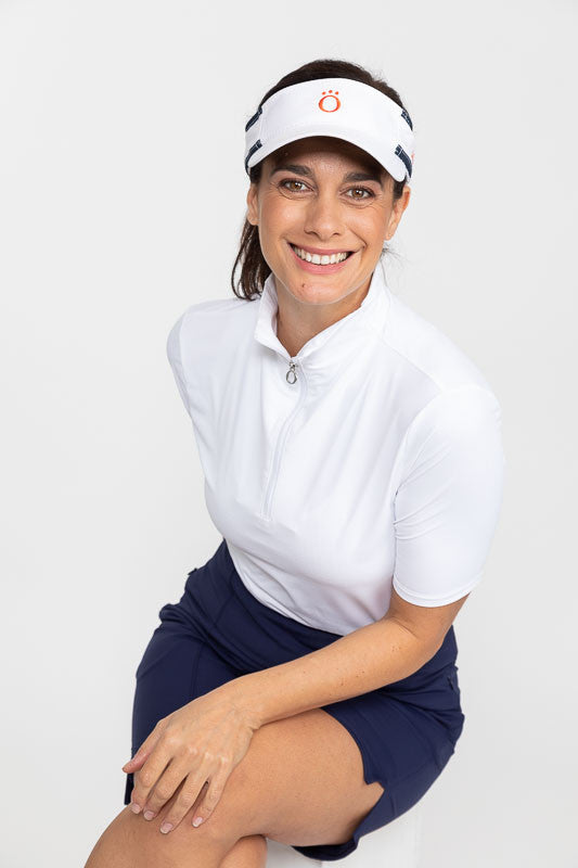 Front view of a seated, smiling woman wearing a white No Hat Hair Visor, a Keep It Covered Short Sleeve Golf Top in white/white, and a Wrap It Up Golf Skort in navy blue