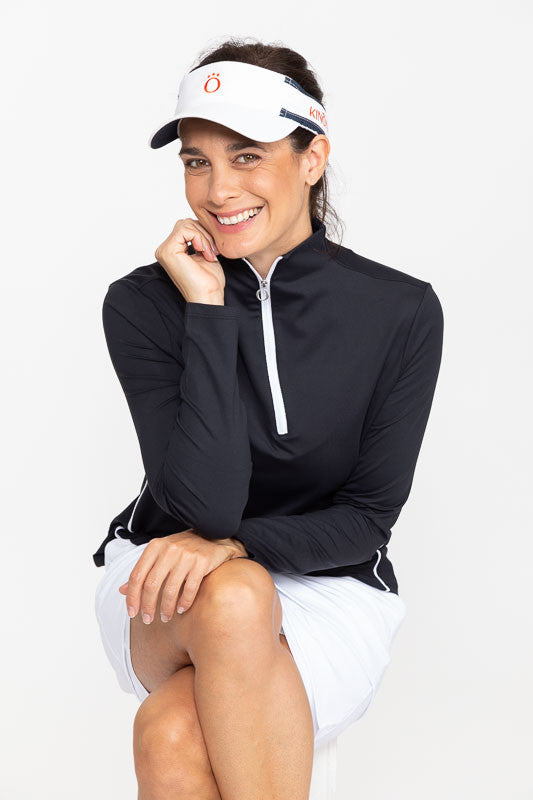 Seated, smiling woman wearing a white No Hat Hair Visor, a Keep It Covered Long Sleeve Golf Top in black, and a Wrap It Up Golf Skort in white