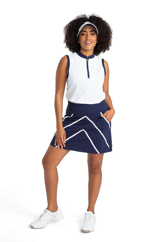 Full front view of a smiling woman wearing the Zip It and Rip It Sleeveless Golf top in white, the Show Me The Way golf skort in navy blue, and a No Hat Hair Visor in white. This top has navy blue accents at the neckline and around each sleeve.
