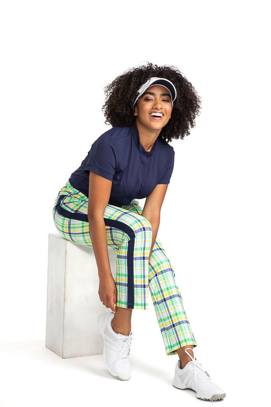 Full front view of a smiling woman sitting and adjusting her pant leg wearing the Band Collar Beauty Short Sleeve Golf Top in Navy Blue, the Smooth Your Waist Crop Pants in Picnic Plaid, and the No Hat Hair Visor in white. 