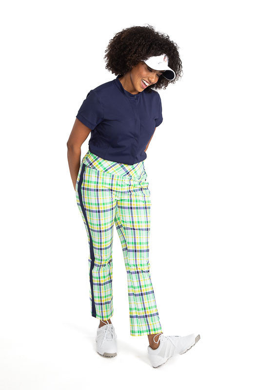 Full front view of a smiling woman wearing the Band Collar Beauty Short Sleeve Golf Top in Navy Blue, the Smooth Your Waist Crop Pants in Picnic Plaid, and the No Hat Hair Visor in white. 