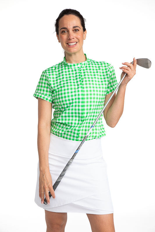 Full front view of a smiling woman holding a golf club across her body wearing the Band Collar Beauty Short Sleeve Golf Shirt in Go Go Gingham and the Wrap It Up Golf Skort in White. The Go Go Gingham print consists of green and white checks.