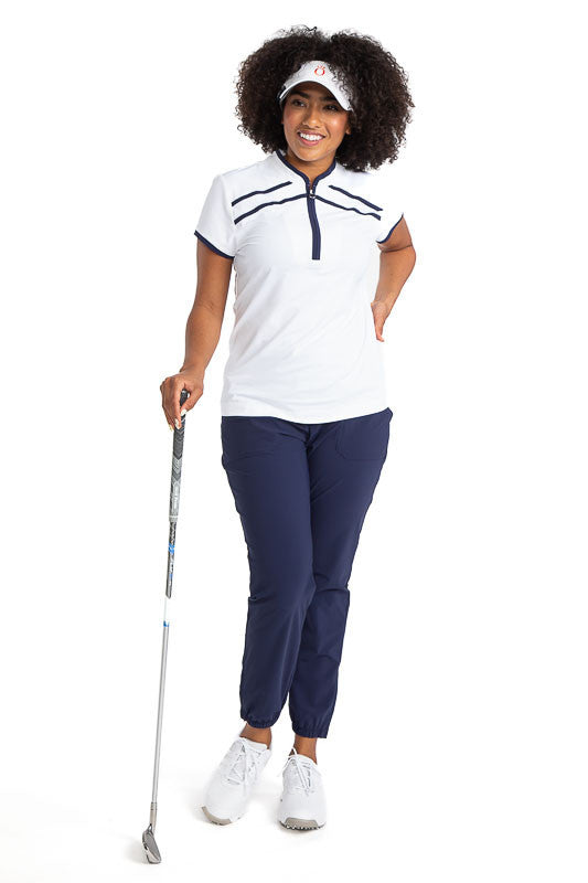 Full front view of a smiling woman golfer wearing the Gimme Putt Short Sleeve Golf Top in White, the Smooth Your Waist Crop Pants in navy blue, and the No Hat Hair Visor in white. This top has navy blue accents along both sides of the zipper, two stripes 