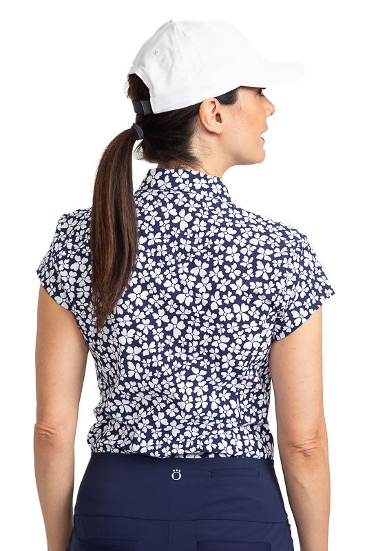 Back view of a woman wearing the Tough in the Rough Short Sleeve Golf Top in Vinca Print and a We've Got You Covered Hat (baseball-style hat) in White. The Vinca print consists of a navy blue background with small, white flowers on it.