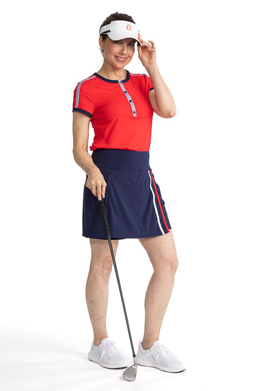Complete front view of a smiling woman golfer wearing the Approach Shot Short Sleeve Golf Top in Cherry Red, the Skort and Short Golf Skort in Navy Blue, and the No Hat Hair Visor in White. This top has navy blue accents around each arm, the neckline, and