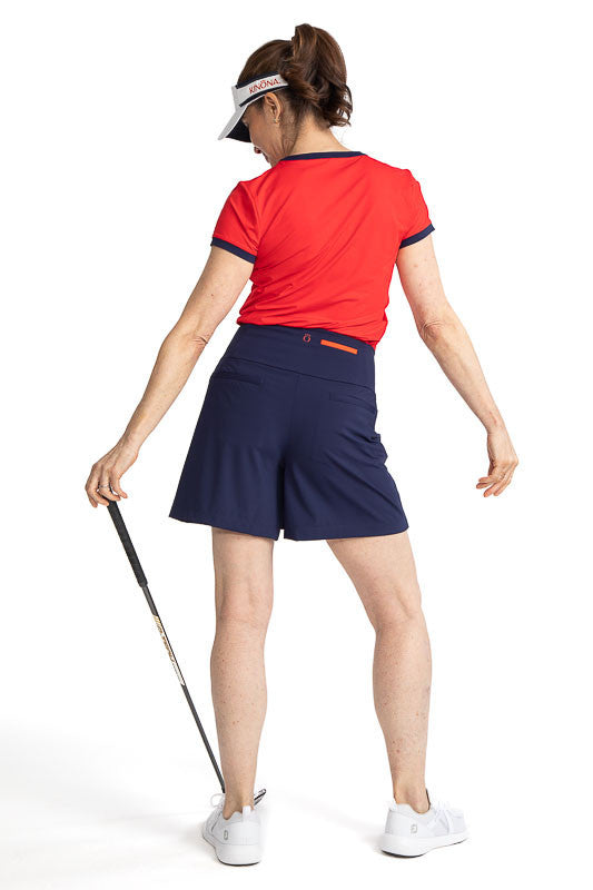 Complete back view of a woman golfer wearing the Approach Shot Short Sleeve Golf Top in Cherry Red, the Skort and Short Golf Skort in Navy Blue, and the No Hat Hair Visor in White. This top has navy blue accents around each arm, the neckline, and the butt