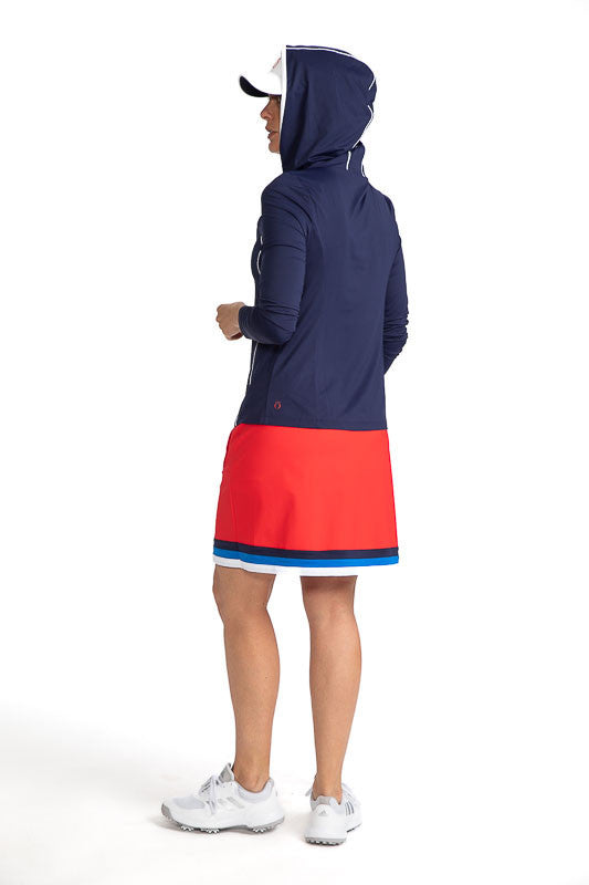 Complete back view of a woman wearing the Layer It Up Long Sleeve Hoodie Golf Top in Navy Blue with the hood pulled up over her No Hat Hair Visor in White, and wearing the On The Fringe Golf Skort in Cherry Red. There are white accents around the edge of 