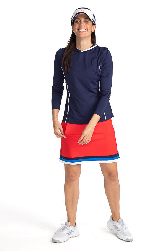 Smiling woman golfer wearing the Layer It Up Long Sleeve Hoodie Golf Top in Navy Blue, the On the Fringe Golf Skort in Cherry Red, and the No Hat Hair Visor in White. There are white accents around the edge of the hood and down each side of this hoodie.
