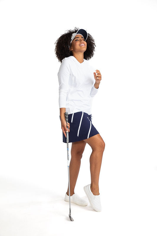 Laughing woman golfer wearing the Layer It Up Long Sleeve Hoodie Golf Top in White, the Show Me The Way Golf Skort in Navy Blue and White, and the No Hat Hair Visor in White.