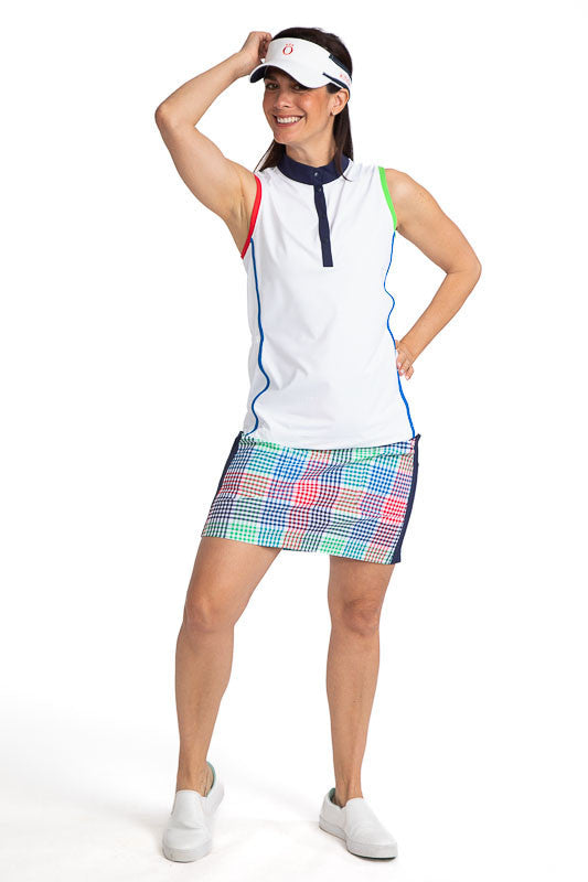 Complete front view of a smiling woman golfer wearing the Pocket Envy Golf Skort in Vacation Plaid, the Make it Snappy Sleeveless Golf Top in White, and the No Hat Hair Visor in White. Vacation Plaid consists of a small, checkered pattern of red, blue, gr