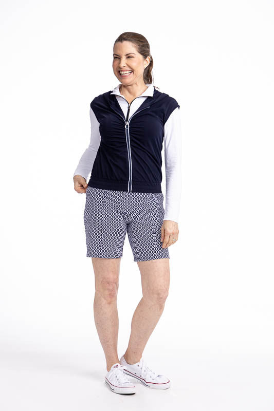Full front view of a smiling woman wearing the Fall Ball Golf Vest in Navy Blue and the Golf Glove Friendly Golf Shorts in Chic Chevron print. This is a solid navy blue vest with a white front zipper and elastic waistband.