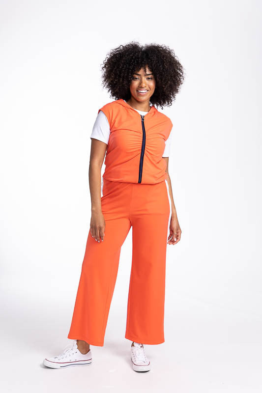 Full front view of a smiling woman wearing the Fall Ball Golf Vest in Coral Red and the Aprés 18 Wide Leg Pants in Coral Red. This is a solid coral red vest with a black front zipper and elastic waistband.