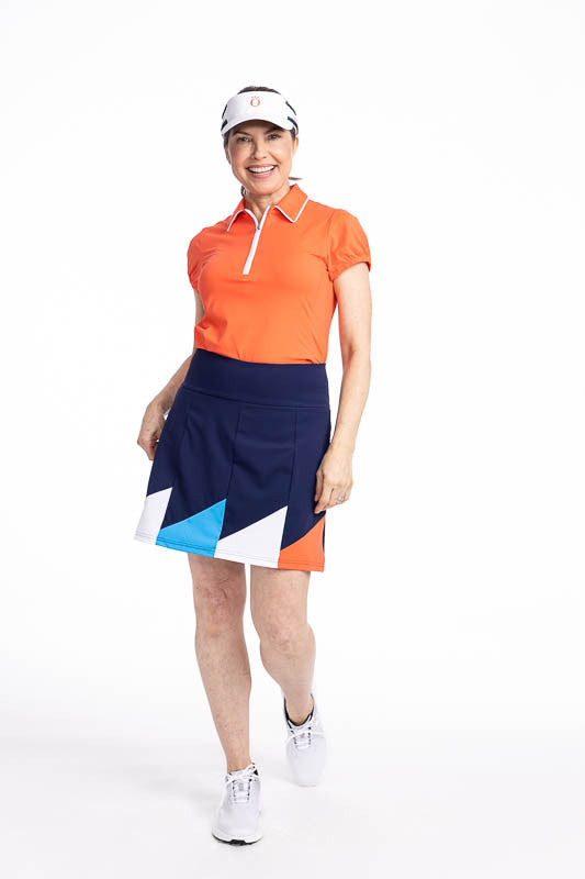 Full front view of a smiling woman wearing the Helping Wind Golf Skort in Navy Blue, the Prettier Than a Polo Short Sleeve Golf Top in Coral Red, and the No Hat Hair Visor in White. This is a navy blue skort with alternating triangles shaped like golf fla