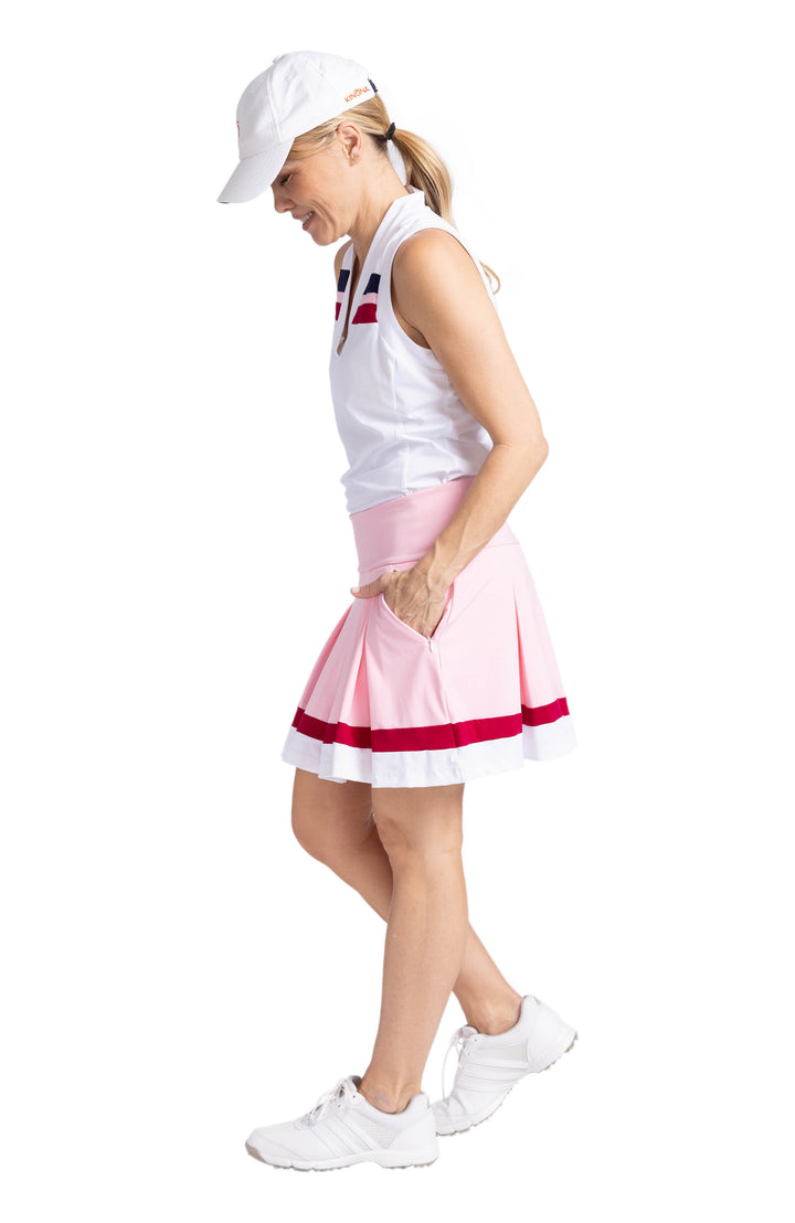 Side view of a woman wearing a white, V-neck, sleeveless top with contrasting navy blue, Bordeaux Red, and white stripes on the upper chest. She is also wearing the Whirl and Twirl Skort in Ballet Pink with her hands in the side pockets of the skort.