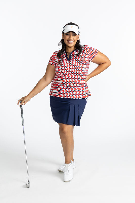 Smiling woman wearing the Party Pleat Golf Skort in Navy Blue, the Classic and Fantastic Short Sleeve Golf Shirt in Chevron Tomato Red, and the No Hat Hair Visor in White. This skort has a solid, white stripe that runs down each side of this skort. 