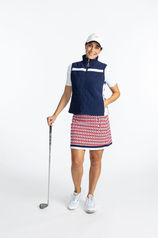 Smiling woman wearing the Chill Layer Golf Vest in Navy Blue, Tee it Up Short Sleeve Golf Shirt in White, the On The Fringe Golf Skort in Chevron Tomato Red, and the We've Got You Covered Hat in White. There is a solid, horizontal white stripe that runs a