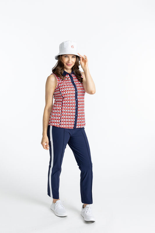 Smiling woman wearing the Dew Sweeper Sleeveless Golf Top in Chevron Tomato Red, the Tailored Track Golf Pants in Navy Blue, and the Big O Bucket Hat in White