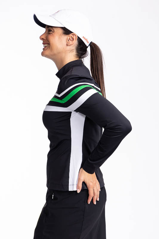 Left side view of a woman wearing the Winter Rules Long Sleeve Golf Top in Black and the We've Got You Covered Hat in White. This top features three alternating stripes of white, rye grass green, and white stripes on a black long sleeve top with one white