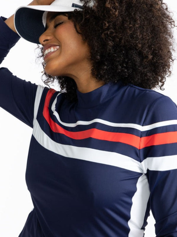Smiling woman wearing the Winter Rules Long Sleeve Golf Top in Navy Blue. This shirt is primarily navy blue with a 360 degree pattern of lines across the chest area. A thin, white stripe followed by a spacer of navy blue, then a thicker, solid tomato red 
