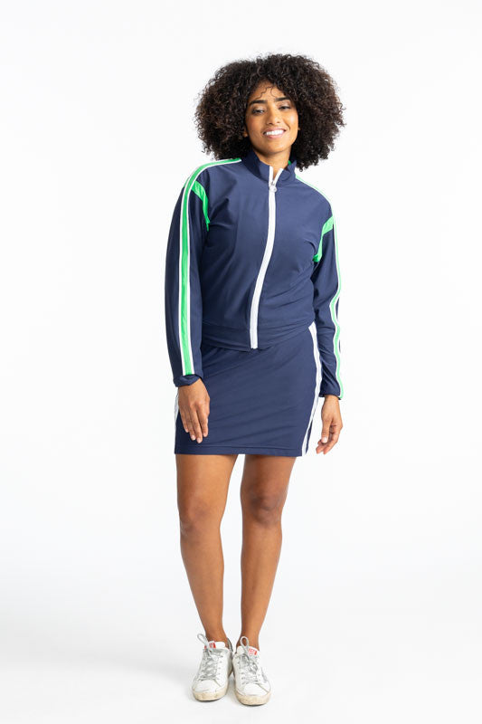 Smiling woman wearing the Warm Up Jacket in Navy Blue and the Seat Warmer Golf Skort in Navy Blue. This jacket is primarily navy blue with a white zipper that runs down the front of this jacket. There is also a Kelly Green stripe that runs around each arm