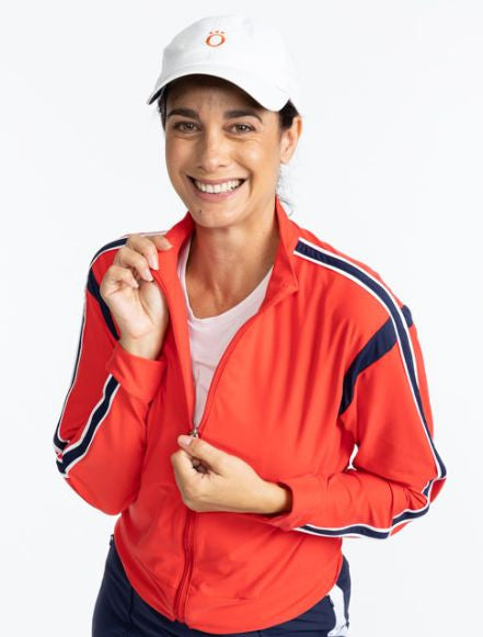 Front view of a smiling woman wearing the Warm Up Jacket in Tomato Red and the We've Got You Covered Hat in White. This jacket is primarily tomato red with a navy blue stripe that runs around each armhole. There is also a navy blue stripe that runs down e