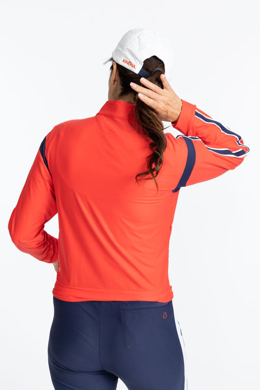 Back view of the Warm Up Jacket in Tomato Red and the We've Got You Covered Hat in White. This jacket is primarily tomato red with a navy blue stripe that runs around each armhole. There is also a navy blue stripe that runs down each sleeve with a thin wh