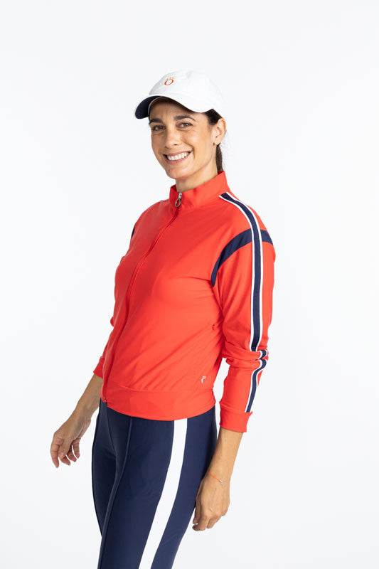Smiling woman wearing the Warm Up Jacket in Tomato Red, the We've Got You Covered Hat in White, and the Ankle Warmer Stirrup Pants in Navy Blue. This jacket is primarily tomato red with a navy blue stripe that runs around each armhole. There is also a nav