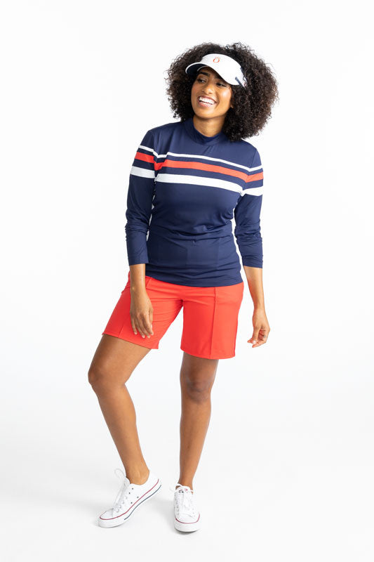 Smiling woman wearing the Golf Glove Friendly Golf Shorts in Tomato Red, the Winter Rules Long Sleeve Golf Top in Navy Blue, and the No Hat Hair Visor in White.