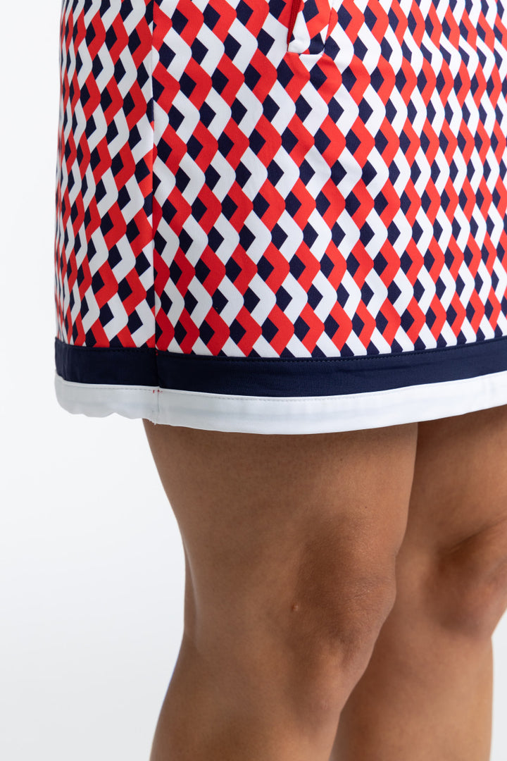 Close right side view of the hemline on the On The Fringe Golf Skort in Chevron Tomato Red. This pattern consists of red, white, and blue chevrons with two solid stripes at the bottom of the skort - a solid navy blue stripe and a solid white stripe.