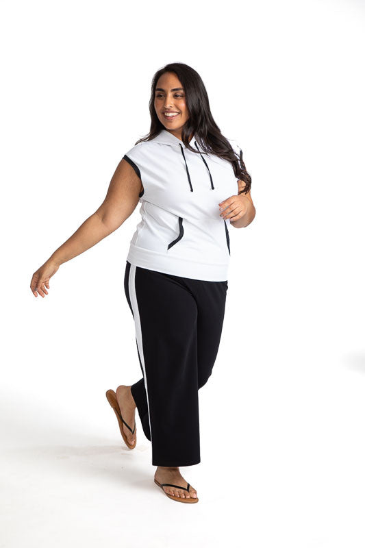 Full front view of a woman golfer wearing the Apres 18 Extended Shoulder Hoodie in White, and the Apres 18 Wide Leg Pants in Black. This hoodie has black accents around each arm hole, black drawstrings at the hoodie, and black accents on each side of the 