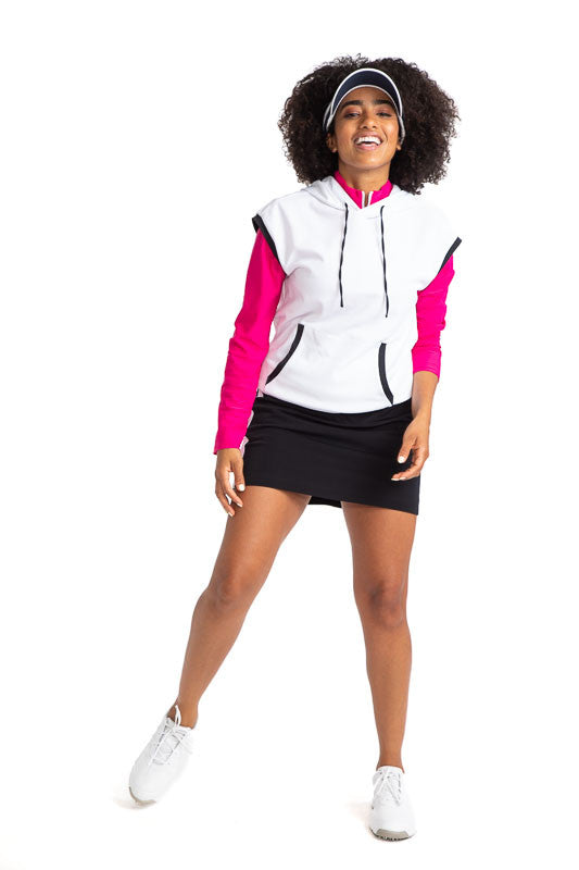 Full front view of a woman golfer wearing the Apres 18 Extended Shoulder Hoodie in White with the Apres 18 Funnel Neck Long Sleeve Top in Magenta Pink, the Apres 18 Sport Skirt in Black, and a No Hat Hair Visor in White. This hoodie has black accents arou