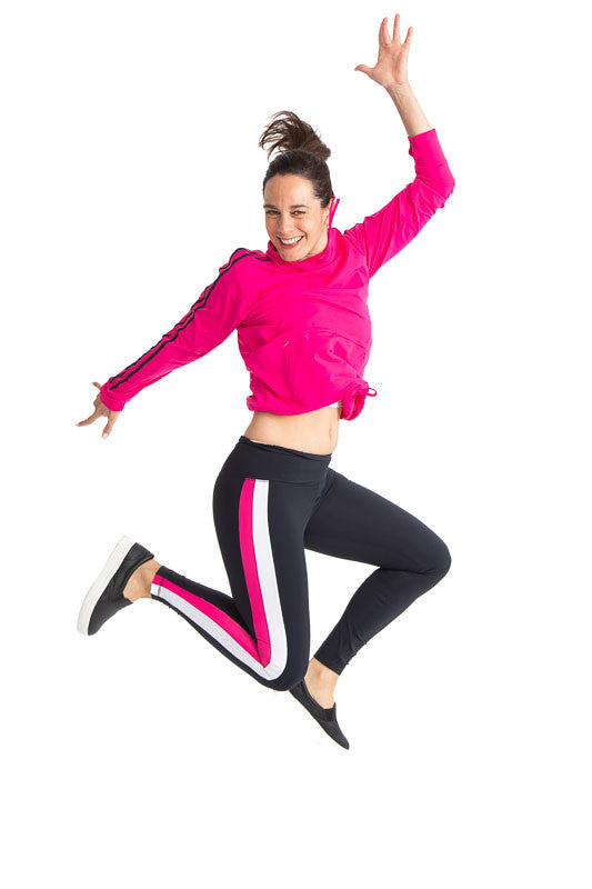 Full front view of a woman jumping in the air wearing the Apres 18 Funnel Neck Long Sleeve Top in Magenta Pink and the Apres 18 Striped Leggings in Black/Magenta. This top has two black stripes running down each arm and magenta pink drawstrings around the