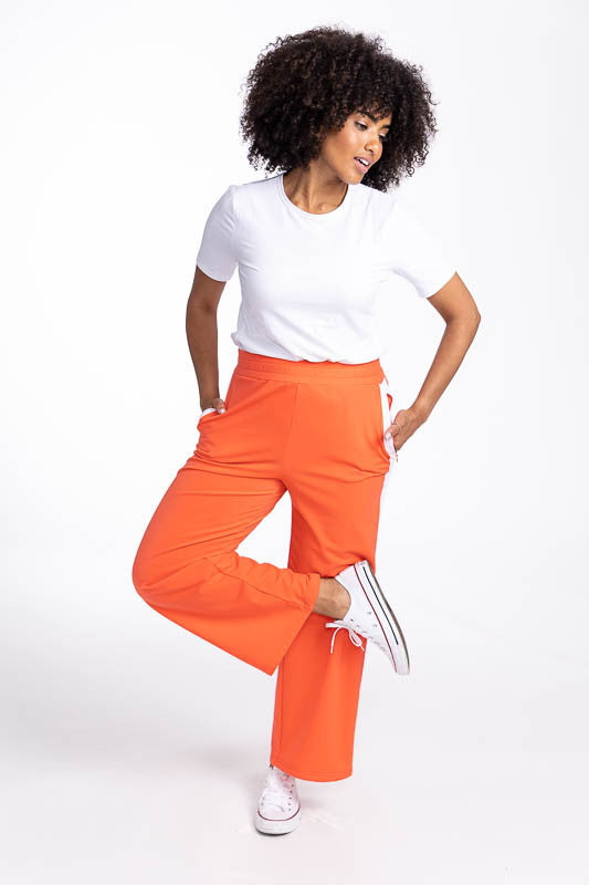 Full front view of a smiling woman wearing the Aprés 18 Wide Leg Pants in Coral Red and the Tee It Up Short Sleeve Golf Top in White. These are a solid coral red pants with one white stripe up each leg.
