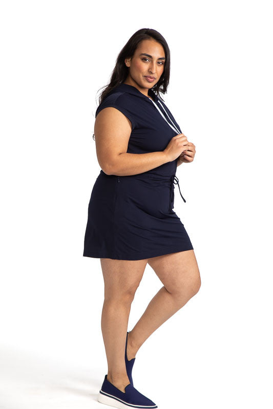 Full right side view of a woman wearing the Aprés 18 Hoodie Dress in navy blue. This dress features a hoodie with adjustable strings for the hood in contrasting white and two front pockets