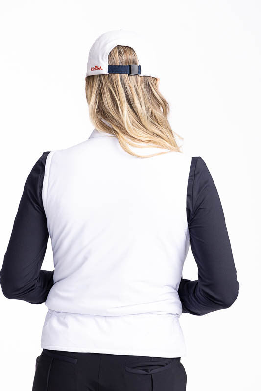 Back view of a smiling woman wearing the Layer Slayer Golf Vest in White, the Lovely Layer Long Sleeve Golf Top in Black, and the We've Got You Covered Hat in White.