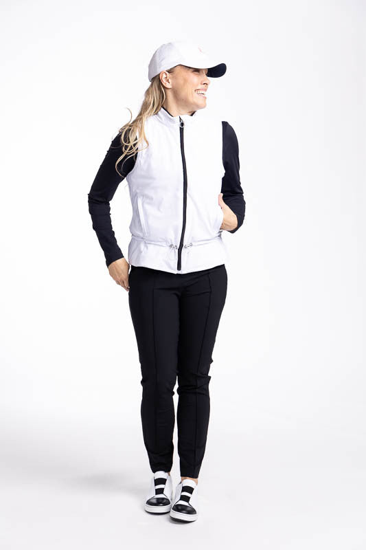 Full front view of a smiling woman wearing the Layer Slayer Golf Vest in White, the Lovely Layer Long Sleeve Golf Top in Black, the Tailored Crop Pants in Black, and the We've Got You Covered Hat in White.