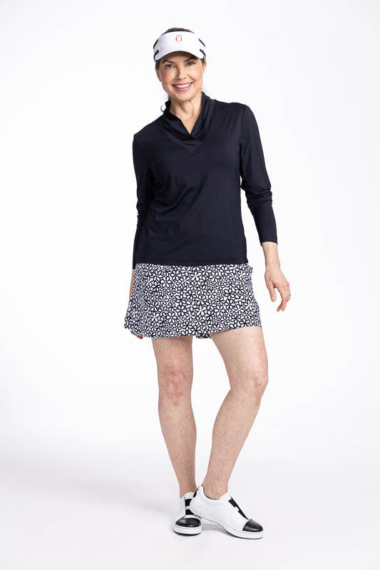 Full front view of a smiling woman wearing the Lovely Layer Long Sleeve Golf Top in Black, the Cool Coulotte Golf Skort in Fall Bloom print, and the No Hat Hair Visor in white.