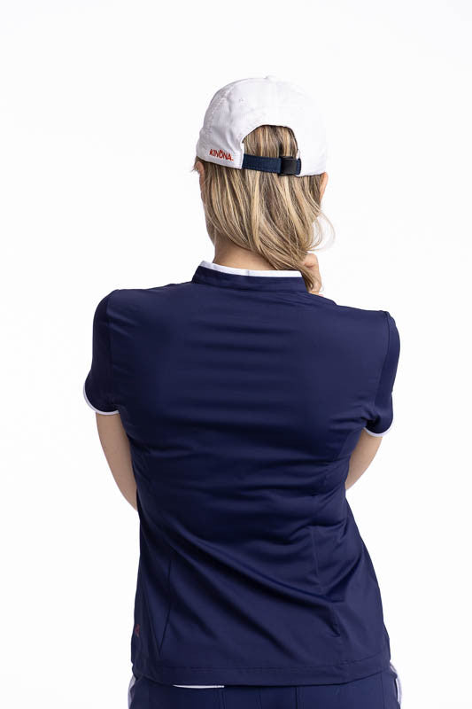 Back view of a smiling woman wearing the Gimme Putt Short Sleeve Golf Top in Navy Blue and the We've Got You Covered Hat in White. The Gimme Putt top is a solid navy blue top with  two diagonal stripes on either side of the front quarter zip - one in cora