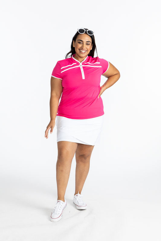 Full front view of a smiling woman wearing sunglasses on her head as well as the Wrap It Up Golf Skort in White/White and the Gimme Putt Short Sleeve Golf Top in Preppy Pink.  This is a solid pink top with a white 1/4 zip front, white trim around the slee
