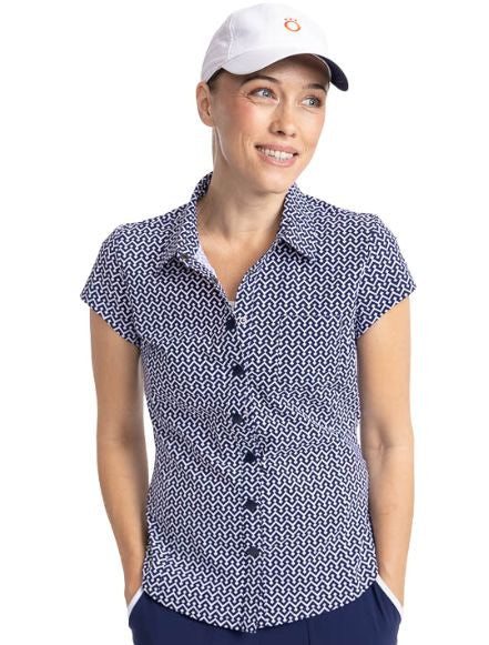 Front view of a smiling woman wearing the Tough in the Rough Short Sleeve Golf Top in Chic Chevron print and the We've Got You Covered Hat in White. The Chic Chevron print consists of white chevrons on a navy blue background. The top features a snap front