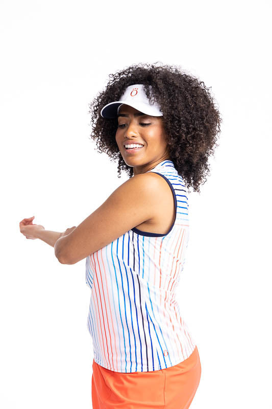 Left side view of a woman wearing the Sandy Par Sleeveless Golf Top in Sun Stripe Print and the No Hat Hair Visor in White. The print consists of a mix of horizontal and vertical stripes in coral red, pacific blue, and navy blue on a white back ground wit
